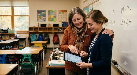 An experienced teacher and a younger colleague reviewing a digital lesson plan on a tablet together in a sunny, quiet elementary school classroom.